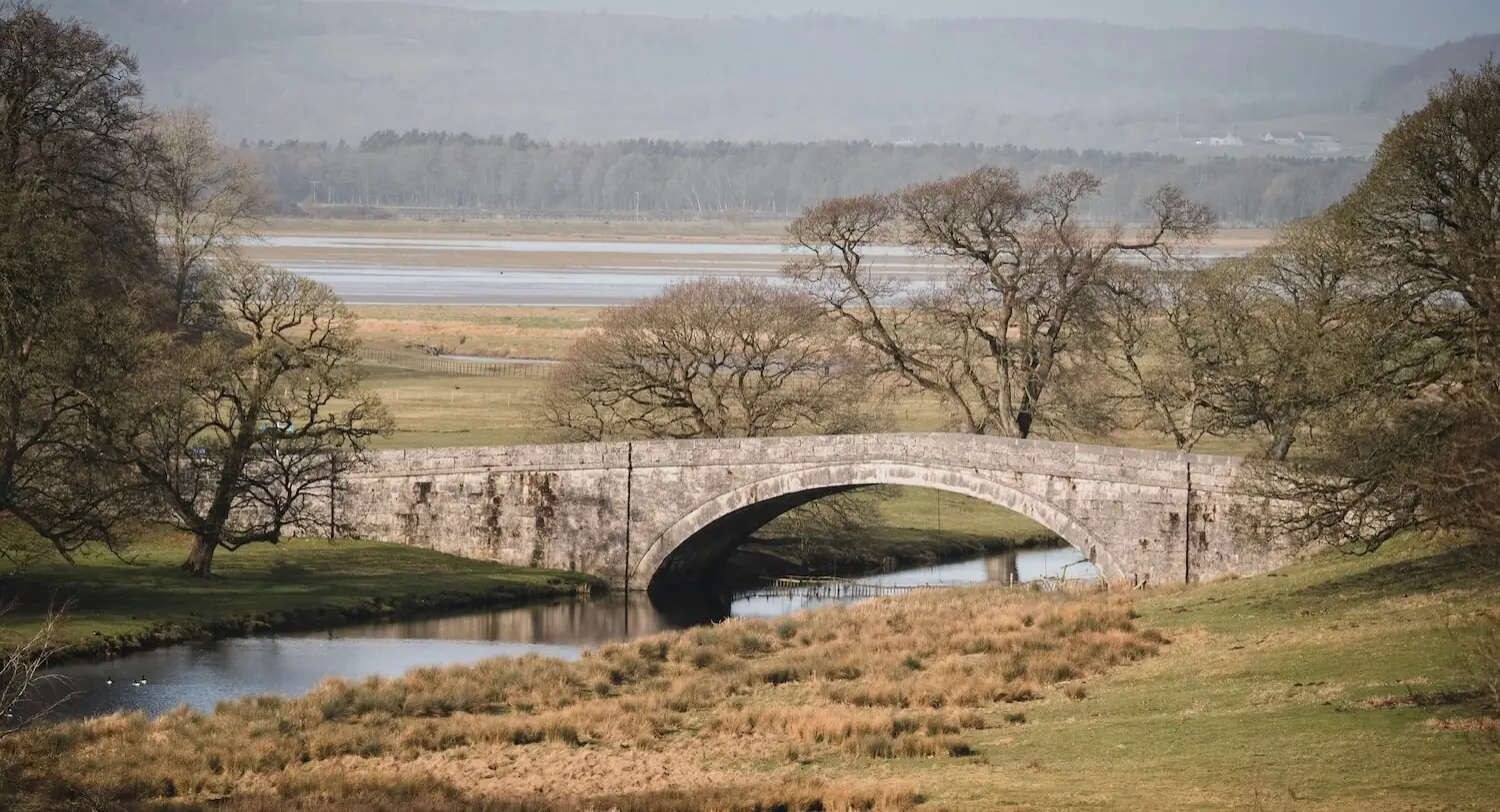 An old single-arch stone bridge over the River Bela in Dallam Deer Park, with the Kent estuary behind