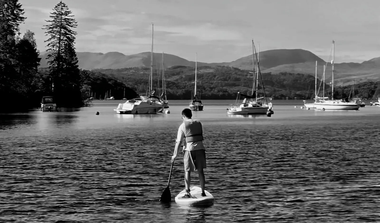 A paddleboarder on Windermere among moored yachts, with the fells behind